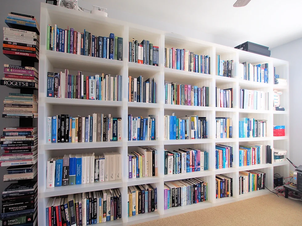 A large white bookshelf filled with various books spans the wall in a bright room. Additional stacks, featuring titles on water treatment, are placed on the left side, next to the shelf. The floor is carpeted, and the room maintains a minimalist and organized appearance.