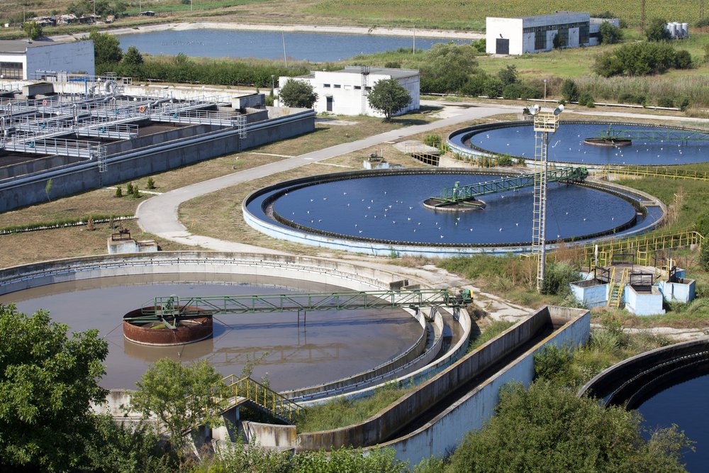 Aerial view of a wastewater treatment plant featuring advanced treatment systems with circular settling tanks and rectangular structures. Surrounded by lush greenery and a body of water, pathways connect different sections, showcasing the integration of emerging technologies in its design.