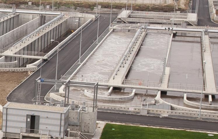 Aerial view of a wastewater treatment plant with multiple rectangular basins filled with water. Concrete structures and pathways divide the areas, showcasing essential processes at work. Machinery is visible, while the foreground reveals a patch of green grass.