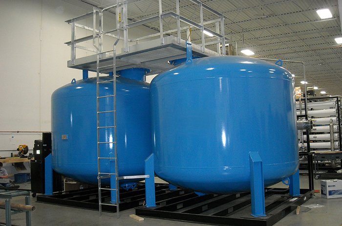 Two large blue industrial tanks with a connected platform and ladder in a warehouse setting.