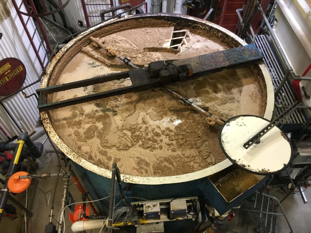 Industrial mixing tank with beige substance, surrounded by pipes and metal framework, viewed from above.