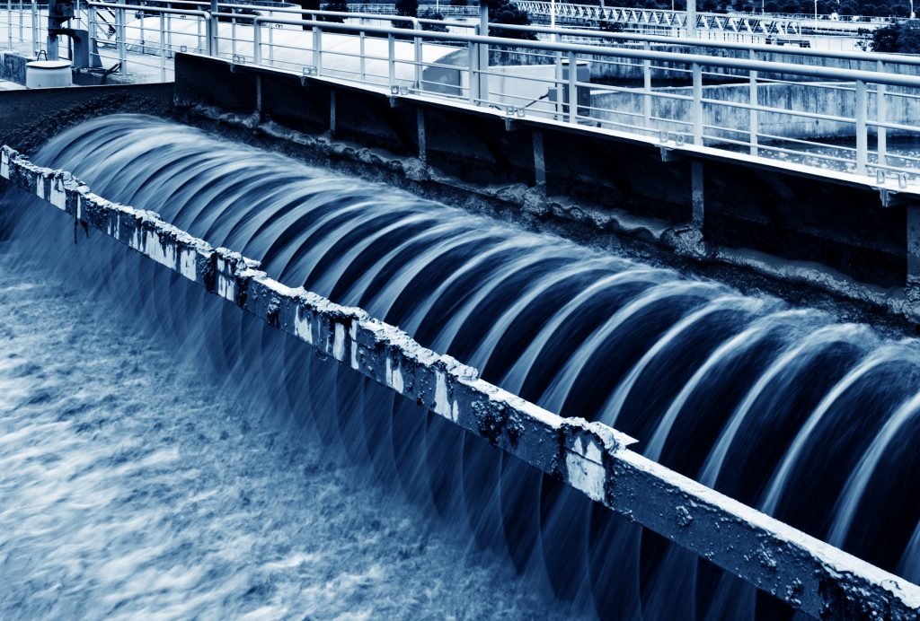 A close-up of a wastewater treatment facility shows water cascading over a series of curved barriers, creating a smooth flow. The scene is tinted in a blue hue, highlighting the structural details and motion of the water, underscoring the facility's treatment efficiency.