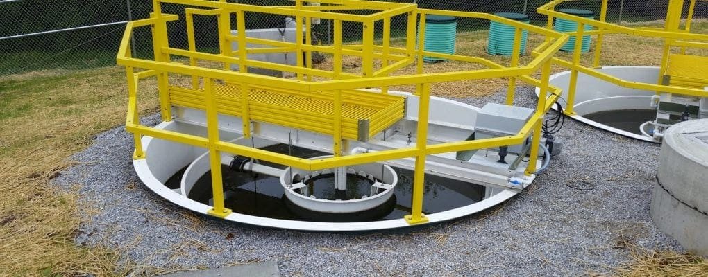 Circular wastewater treatment tank with yellow safety railings, surrounded by gravel and grass, under a clear sky.