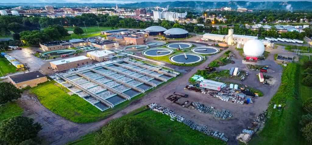 Aerial view of a wastewater treatment plant with settling tanks and various industrial buildings surrounded by green areas.