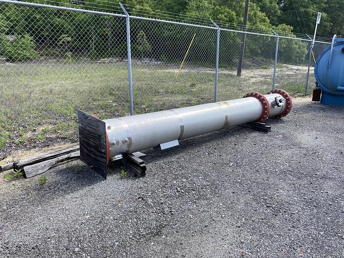 A long, cylindrical metal pipe with flange ends, resembling a packed column used in water treatment, is placed horizontally on wooden supports on a gravel surface. It sits beside a chain-link fence with greenery and trees in the background, highlighting the efficiency of its installation.