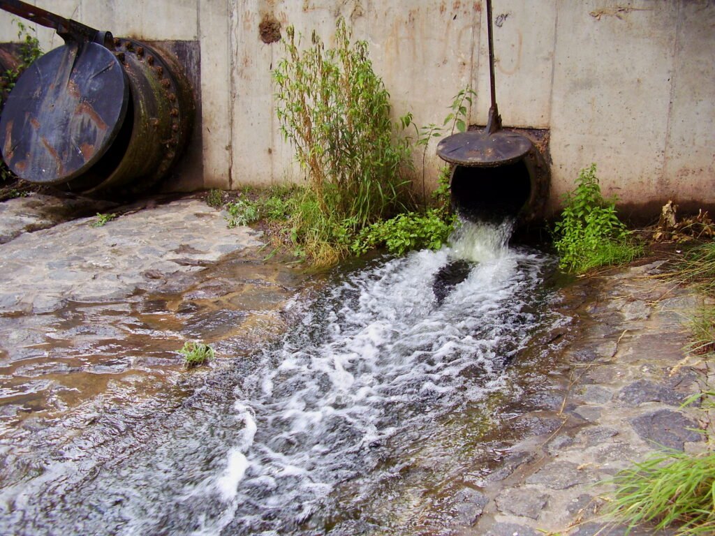 Water flowing from a drainage pipe into a stone-lined channel, surrounded by plants and a concrete wall.