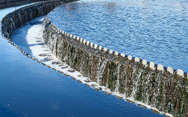 A curved weir, integral to wastewater treatment, forms a gentle cascade that separates a body of water. The water uniformly flows over the weir's edge, creating small waterfalls with white froth at the base against a blue background.