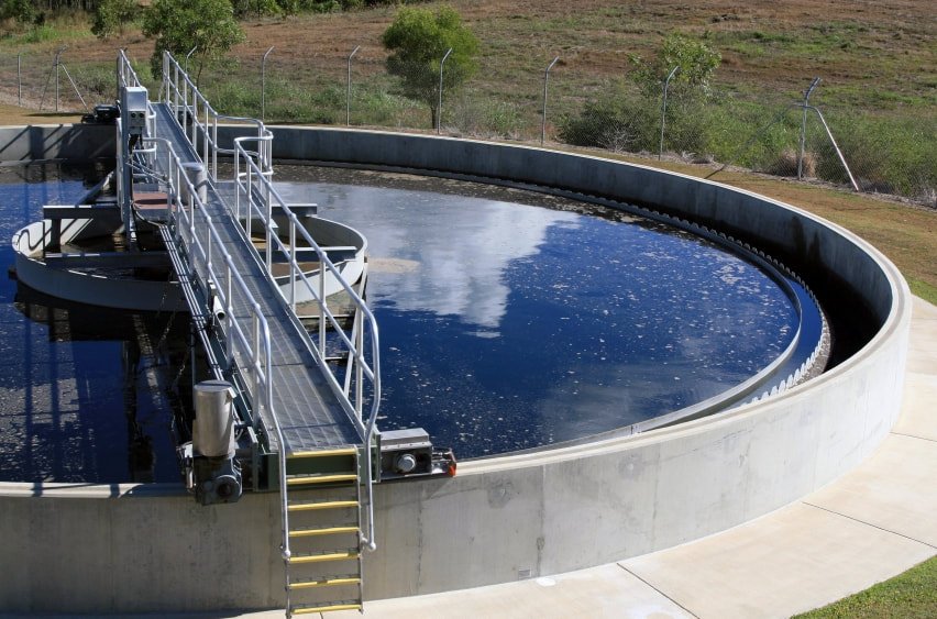 A circular wastewater treatment tank with a metal walkway above it is filled with dark water. The background features a grassy area and a tree, with a fence encircling the facility. The sky above is reflected in the water's surface, indicating careful secondary clarification to maintain effluent quality.