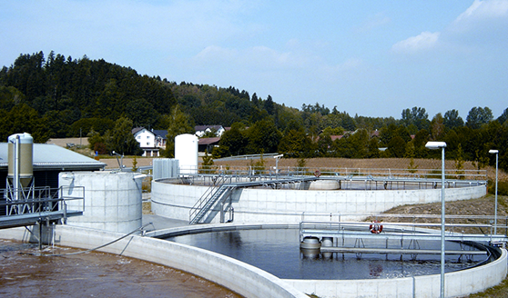 A wastewater treatment facility employs secondary treatment with circular concrete structures, filled with water, nestled in a rural area with trees and a few buildings in the background under a blue sky.