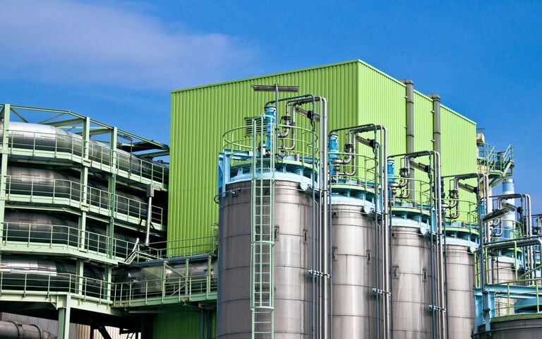 An industrial plant featuring metal silos and a bright green building, equipped for wastewater treatment. Numerous pipes and walkways intertwine, offering process insights under a clear blue sky.