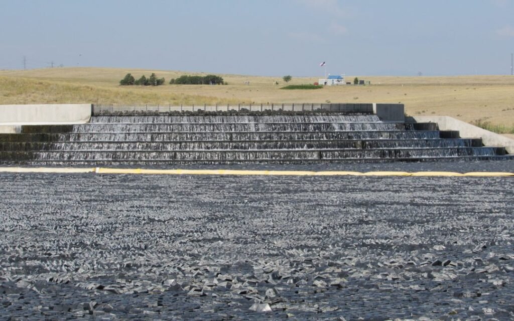 A stepped spillway with flowing water leads to a large reservoir filled with black plastic shade balls. In the background, under a clear sky, the growing city of Aurora's Prairie Waters Project thrives, with a distant building perched on a grassy hill.