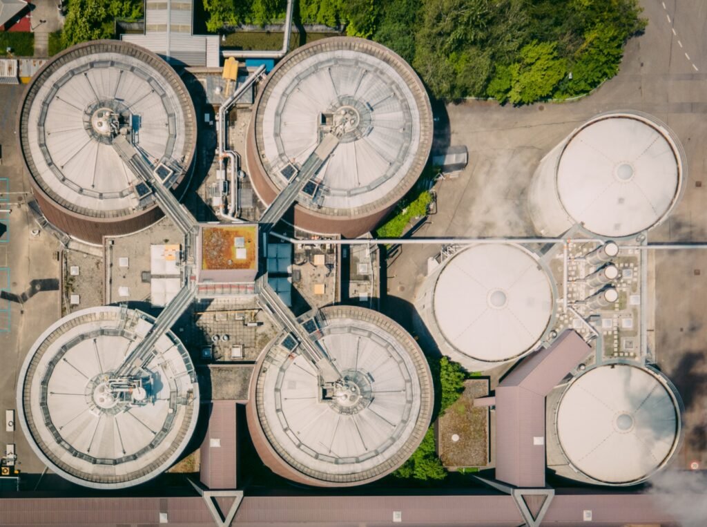 Aerial view of a wastewater treatment facility featuring four large circular tanks connected by a cross-shaped walkway. Adjacent are four smaller cylindrical tanks. Green trees border part of the complex, while concrete and metal structures surround the area.