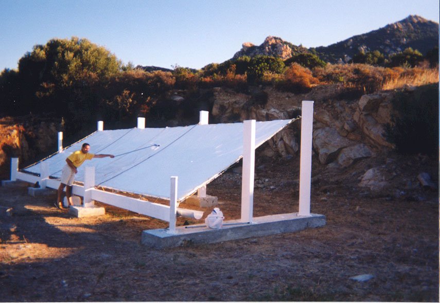 A person in a yellow shirt is adjusting a large, white, slanted solar panel structure outdoors. Supported by wooden beams on grassy terrain, it integrates dew collection systems amid rocky hills and trees under a clear sky.