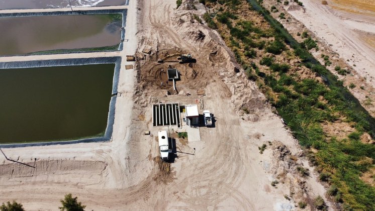 Aerial view of a hybrid construction site featuring vehicles, equipment, and partially built structures near a series of rectangular constructed wetlands. The surrounding area is arid, with sparse vegetation and sandy terrain.