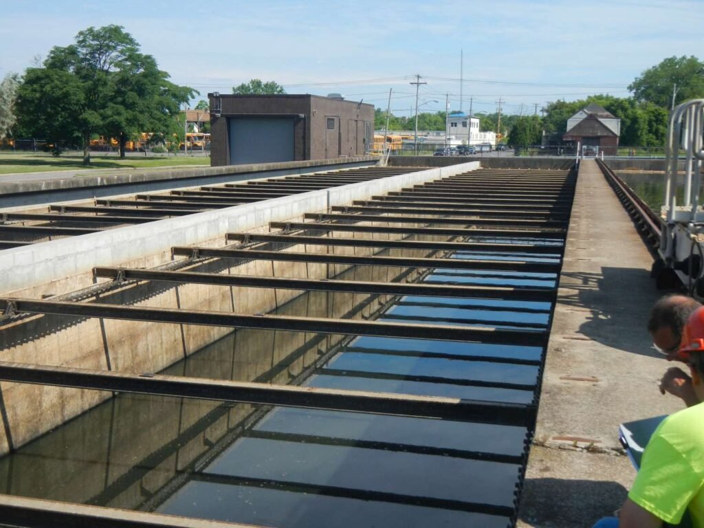 The image depicts a wastewater treatment plant with large rectangular tanks and metal beams. A small building, trees, and utility poles are in the background. In the foreground, a person in a red shirt relaxes on a concrete ledge, evoking a scene reminiscent of Niagara Falls.