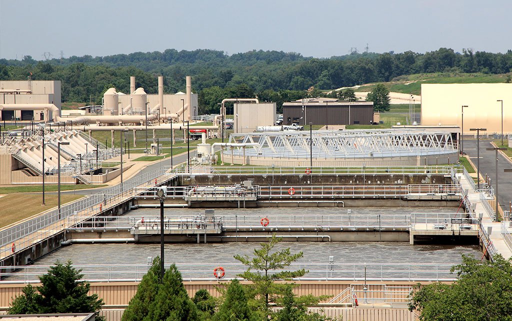 Aerial view of the Upper Occoquan Service Authority's Regional Water Reclamation Plant, featuring large rectangular tanks and industrial structures. Trees and green areas surround the facility, with distant forested hills under a clear sky. Orange life rings are visible along the tanks.