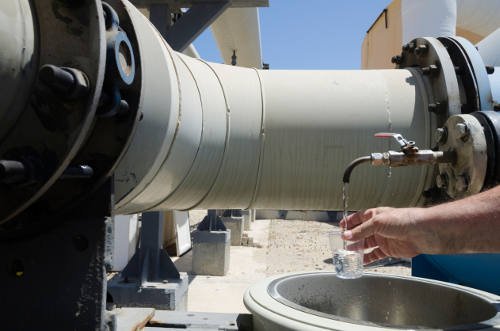 A large industrial pipe with a faucet, possibly part of a desalination process, fills a small glass with water. A person holds the glass under the running water. In the background, metal structures rise against a clear blue sky.