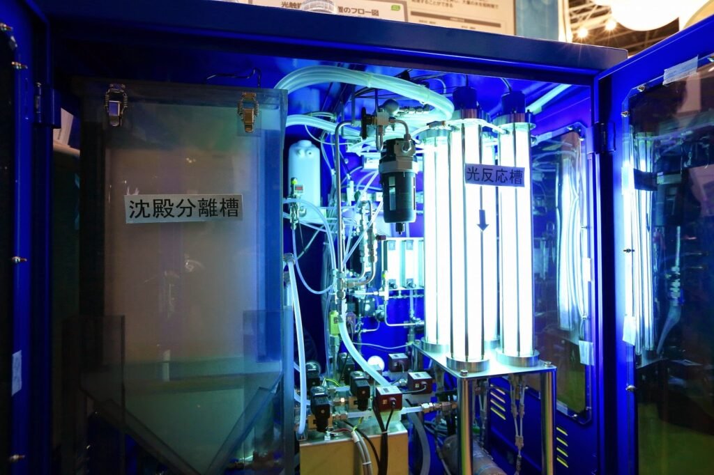 A complex machine in a blue enclosure features various tubes, wires, and illuminated components for water purification. Japanese text labels parts of the machine, including a tall narrow tank with blue lights and clear containers designed for photocatalytic processes.