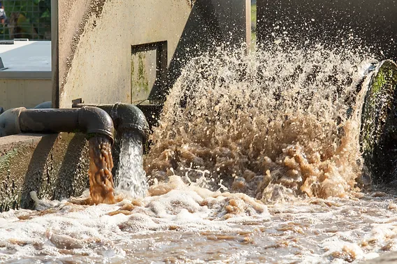 Wastewater flows vigorously from two pipes into the treatment facility, showcasing the activated sludge process. Splashes and frothy waves emerge as the water, brownish yet essential for testing, swirls around. Concrete structures stand in the background, highlighting efficient wastewater treatment underway.