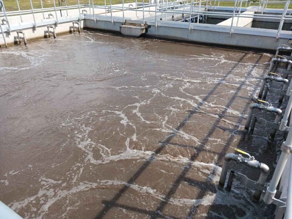 A wastewater treatment facility features a large aeration basin filled with swirling, aerated water. Pipes and valves line the edges, and railings surround the area. The surface shows frothy bubbles and movement, illustrating an essential part of effective wastewater treatment.