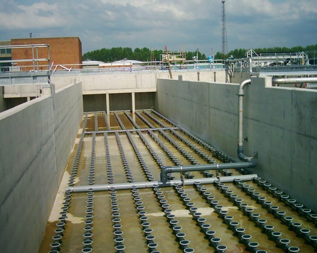 A large concrete structure with numerous circular discs evenly lined on the floor, pipes running along the bottom and sides, houses an essential aeration blower system in an industrial setting, with a cloudy sky forming a dramatic backdrop.