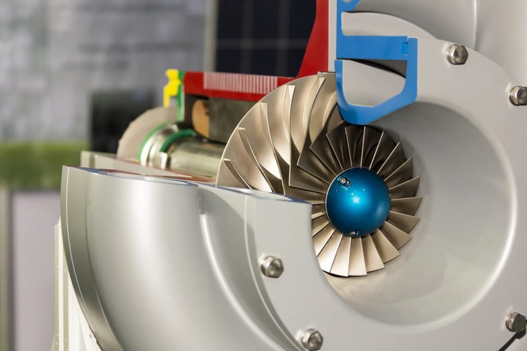 Close-up of a turbine engine component featuring a metal rotor with curved blades, akin to advanced aeration blowers. The blades encircle a central blue hub, surrounded by a gray housing with bolts. The background is blurred, emphasizing the engineered detail of the machinery.