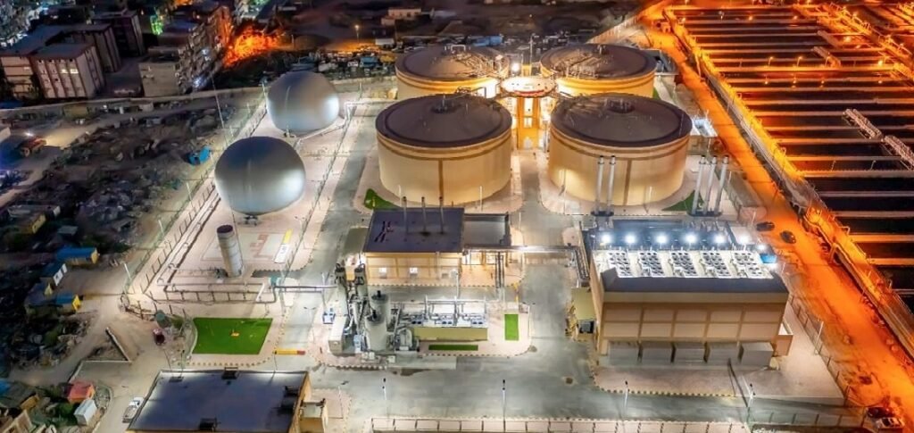 Aerial view of the Alexandria Wastewater Treatment Plant at night, showcasing well-lit industrial spaces. The scene features large cylindrical and dome-shaped structures, with surrounding buildings illuminated and pathways leading to visible parking areas, painting a striking urban landscape.