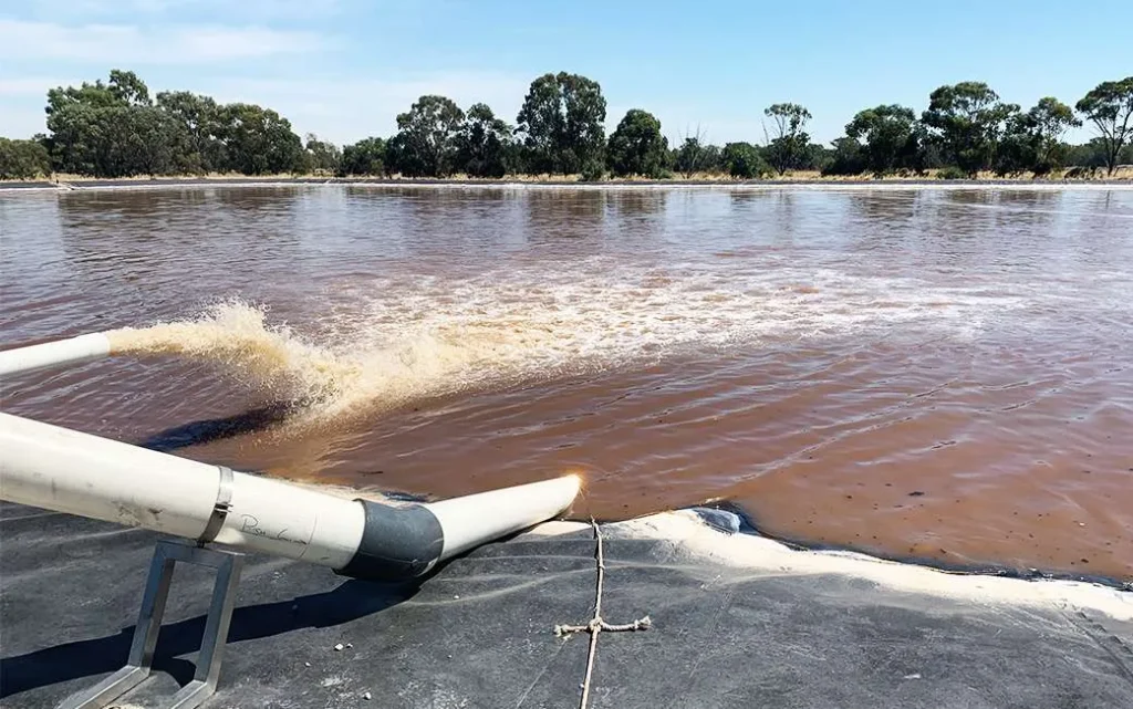 A large pipe discharges wastewater into a body of brownish water surrounded by trees under a clear blue sky. The water splashes as it exits the pipe, and a black liner is visible on the ground near the pipe.