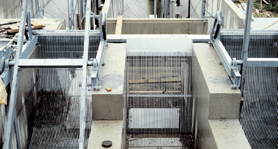 Metal bar screens in a water treatment facility help filter debris, surrounded by concrete infrastructure and greenery.