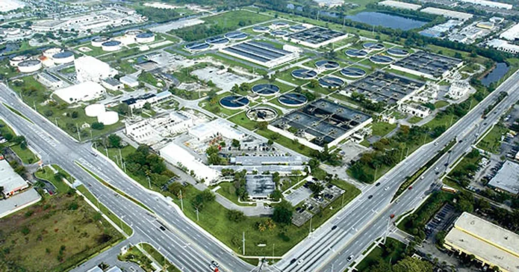 An aerial view reveals a regional wastewater treatment complex, featuring numerous circular tanks and rectangular buildings. Nestled in Broward County, the facility is surrounded by roads with vehicles, green spaces, and a few smaller structures.