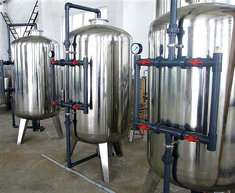 Three large, shiny stainless steel tanks, essential for carbon adsorption in wastewater treatment, are connected by black and red valves, standing on metal supports in an industrial setting. A pressure gauge is visible on one tank. A ladder is propped against the white wall in the background.