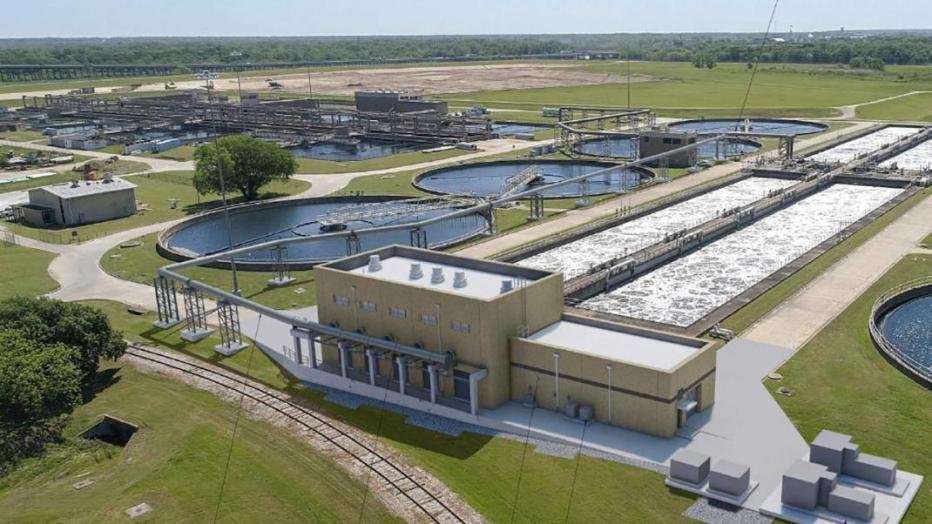 Aerial view of the Central Wastewater Treatment Plant near Dallas, showcasing circular and rectangular tanks amidst lush green fields. A beige industrial building stands prominently in the foreground, set against a backdrop of open land and distant trees.