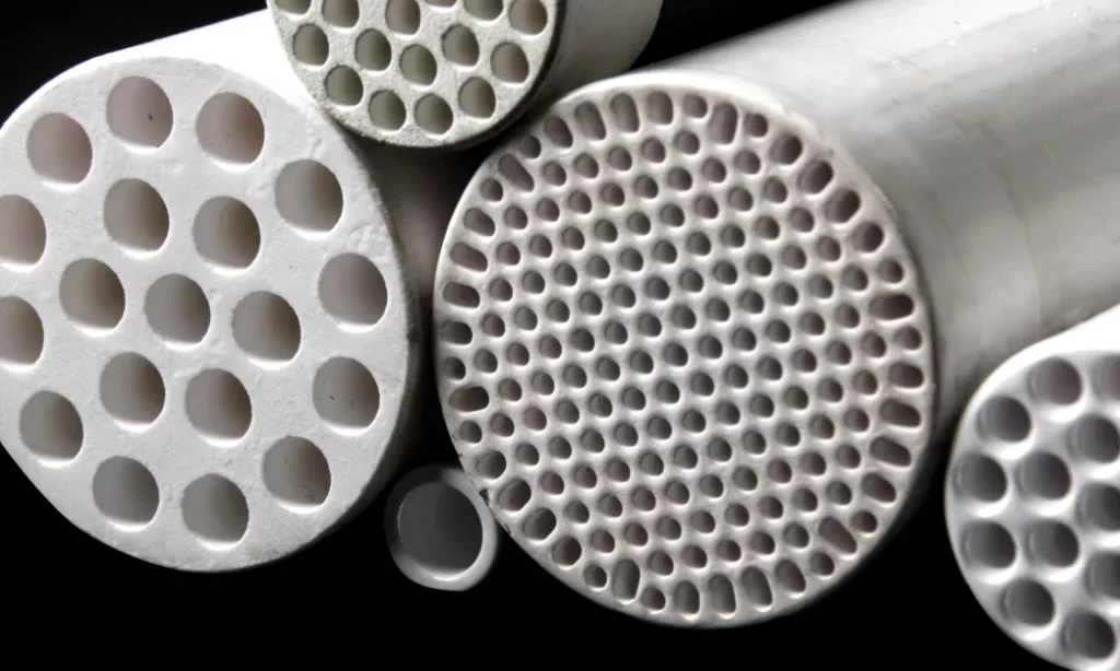 Close-up of several white ceramic membrane tubes with various configurations of round holes. The tubes, part of a filtration system, are arranged in a cluster against a dark background, showcasing different patterns and sizes of perforations.