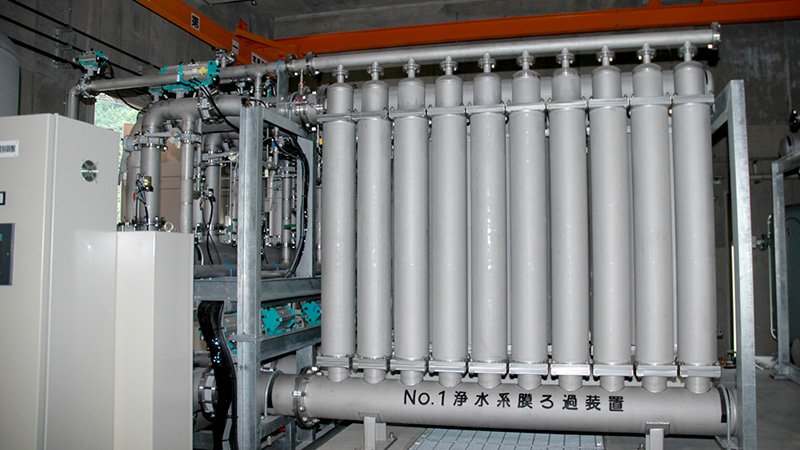 Industrial water purification equipment featuring a system of vertical cylindrical filters and interconnected pipes, labeled with Japanese text. The setup utilizes advanced membrane filtration technology, including various valves and gauges, in a utility room with concrete walls.