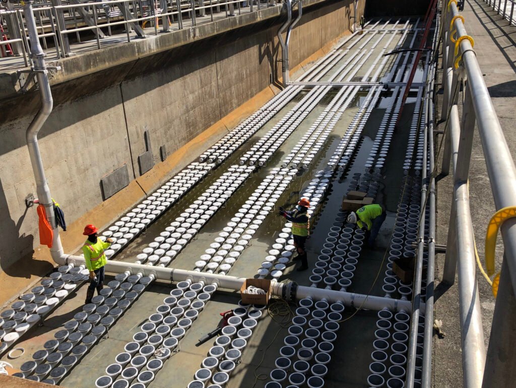 Workers in safety gear are diligently installing or maintaining multiple white circular devices in a large, empty concrete water channel at a Charlotte Water treatment plant. The setup appears organized in rows, and sunlight illuminates the area. Safety railings are visible along the perimeter.