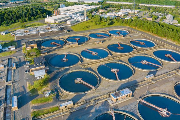 Aerial view of a wastewater treatment plant showcasing efficient distribution strategies with multiple large circular tanks. The facility, surrounded by greenery and nearby industrial buildings, features blue tanks interconnected by walkways and infrastructure for optimal reach.