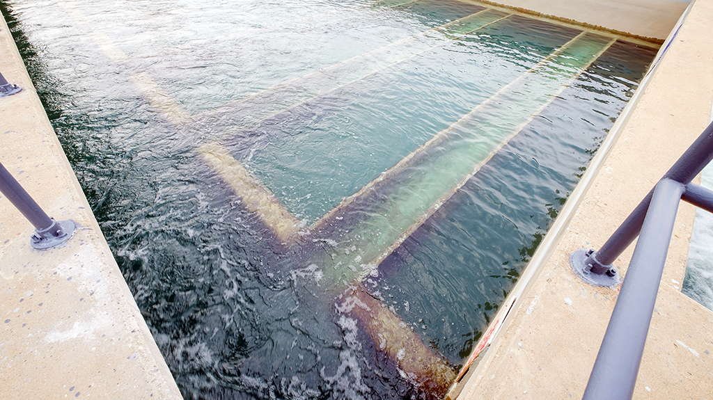 A rectangular, concrete-lined water channel filled with flowing water reveals gentle ripples on its surface. Submerged metal beams create linear patterns, hinting at simple technology in use. Metal railings line the edges of this gravity-based filtration system, ensuring enhanced water purity.
.