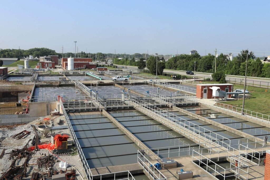 Aerial view of the Belmont Advanced Wastewater Treatment Plant in Indianapolis, showcasing multiple rectangular pools, metal railings, and buildings. Roads and greenery surround the facility under a clear blue sky.