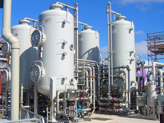 An industrial scene showcases multiple large vertical metal tanks connected by a network of pipes and valves, part of an advanced water purification facility. Under a clear blue sky, scaffolding is visible on the right, highlighting the ongoing ion exchange upgrades.