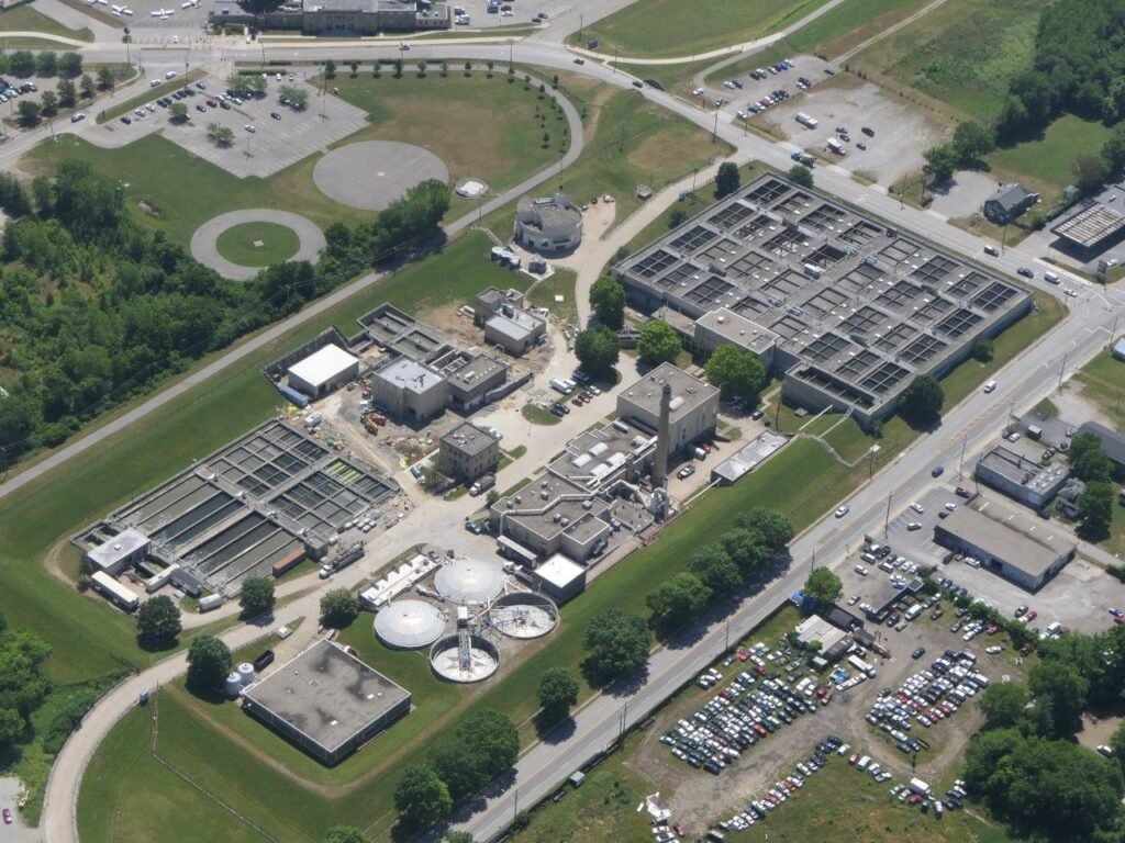 Aerial view of the Lemay Wastewater Treatment Plant, showcasing multiple rectangular tanks and circular structures. The facility is surrounded by roads, grassy areas, and a parking lot filled with cars. Nearby are open fields and scattered trees.