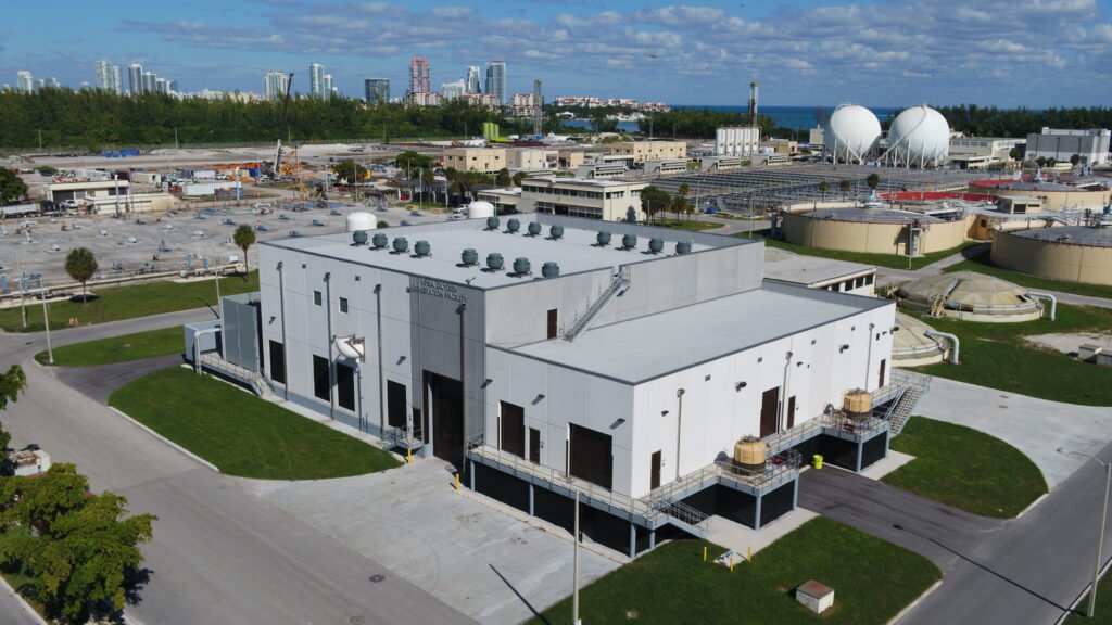 An aerial view captures a large industrial facility, likely a wastewater treatment facility, with a flat roof and HVAC units. Surrounding structures include open land areas and water tanks. In the distance, high-rise buildings rise against a blue sky dotted with clouds.