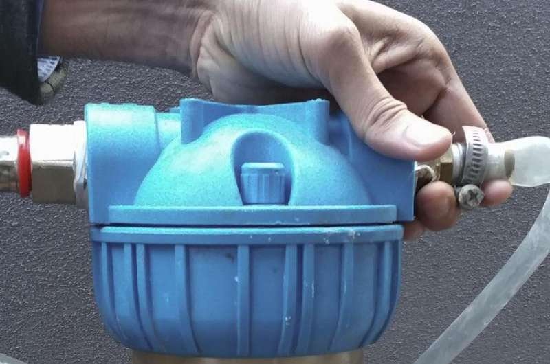 A hand adjusts a blue plastic water filter, integral in nanocellulose-based purification, installed against a dark background. The filter features tubular connections on each side, with the hand gripping a valve or knob on the upper part.