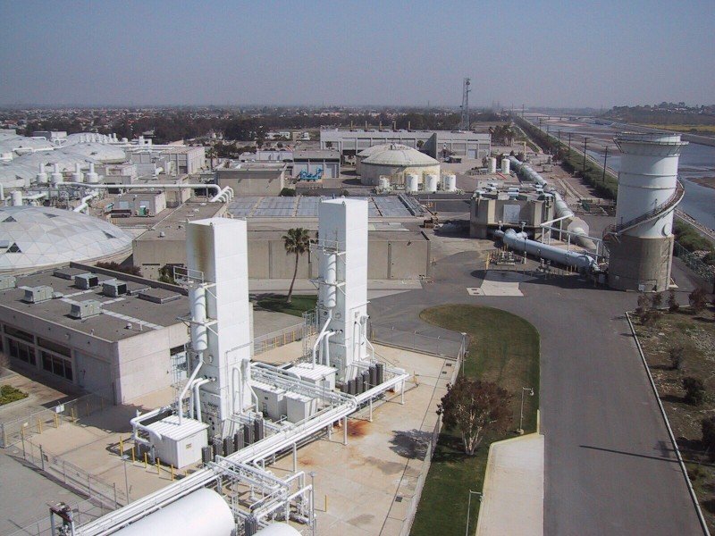 An aerial view of the expansive Orange County Sanitation District's Plant No. 2 showcases a busy industrial facility with multiple buildings and structures, including towering pipes and tanks integral to its wastewater treatment process. The site is bordered by roads and a canal under clear skies.