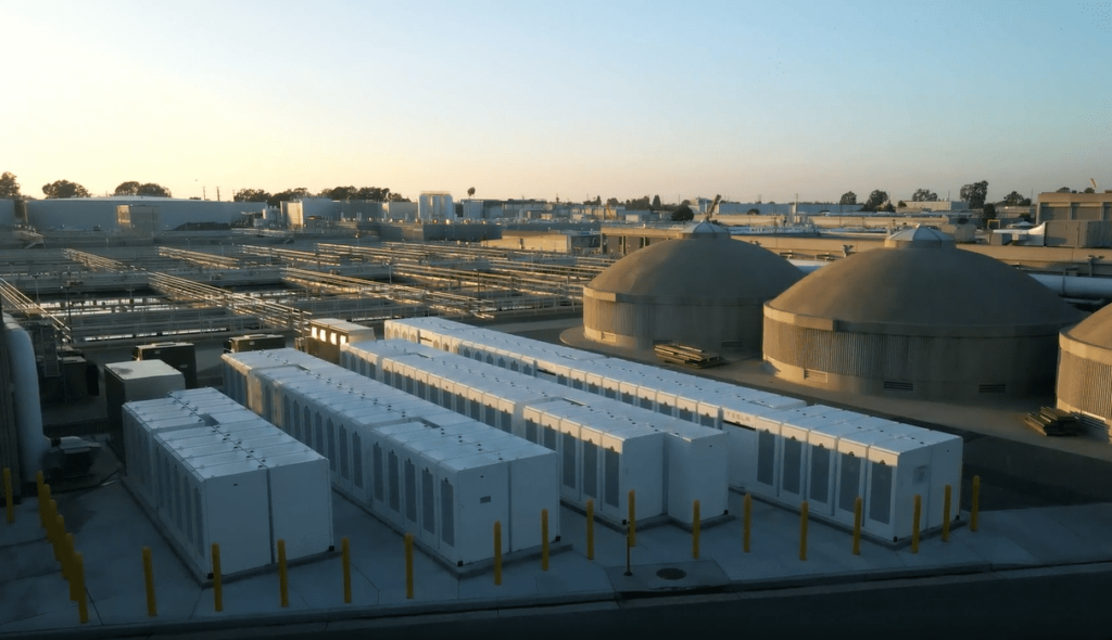 Industrial facility at Plant No. 1 with large tanks and numerous white rectangular units. Pipes run across the area, bathed in warm, late-afternoon sunlight. Trees and additional structures of the Orange County Sanitation District are visible in the background.