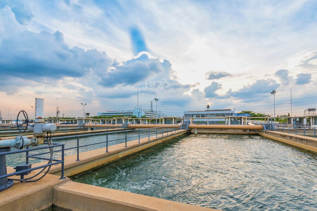 A water treatment facility under a cloudy sky features large rectangular basins used for ozonation, an advanced water purification method. Surrounding railings and pipes connect the system to buildings and industrial structures visible in the background.