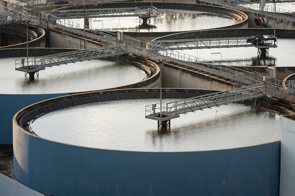 Aerial view of circular water treatment tanks at an industrial plant highlights sustainable management. The tanks, with metal walkways on top, reflect calm waters amidst concrete barriers and metal structures, showcasing a strategic approach to industrial water management.