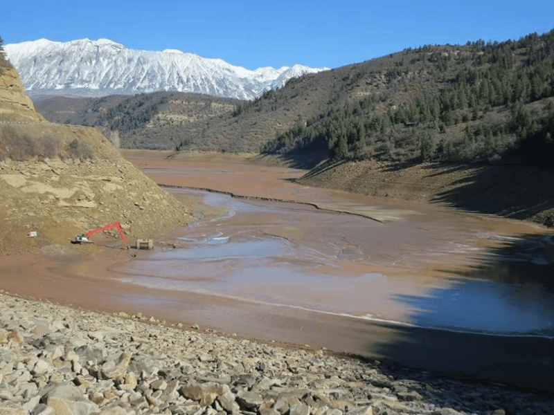 Dry lakebed with construction equipment, surrounded by mountains and trees under a clear blue sky.