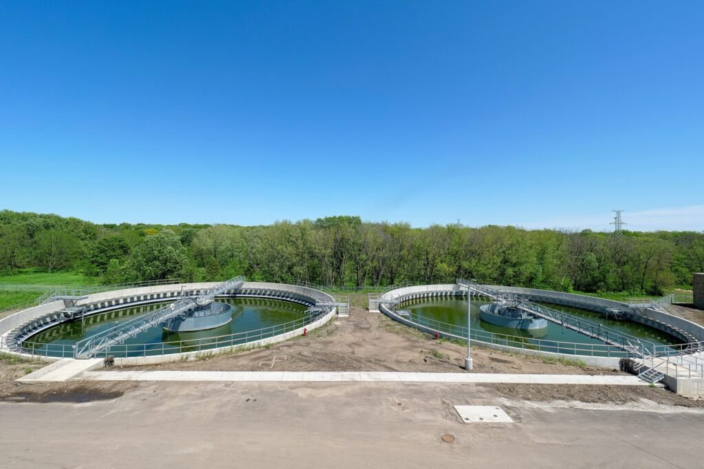 Aerial view of two circular wastewater treatment tanks at the South River plant, surrounded by lush greenery. Clear blue sky above, with trees in the background. Concrete paths and fences encircle the tanks, holding dark green water.