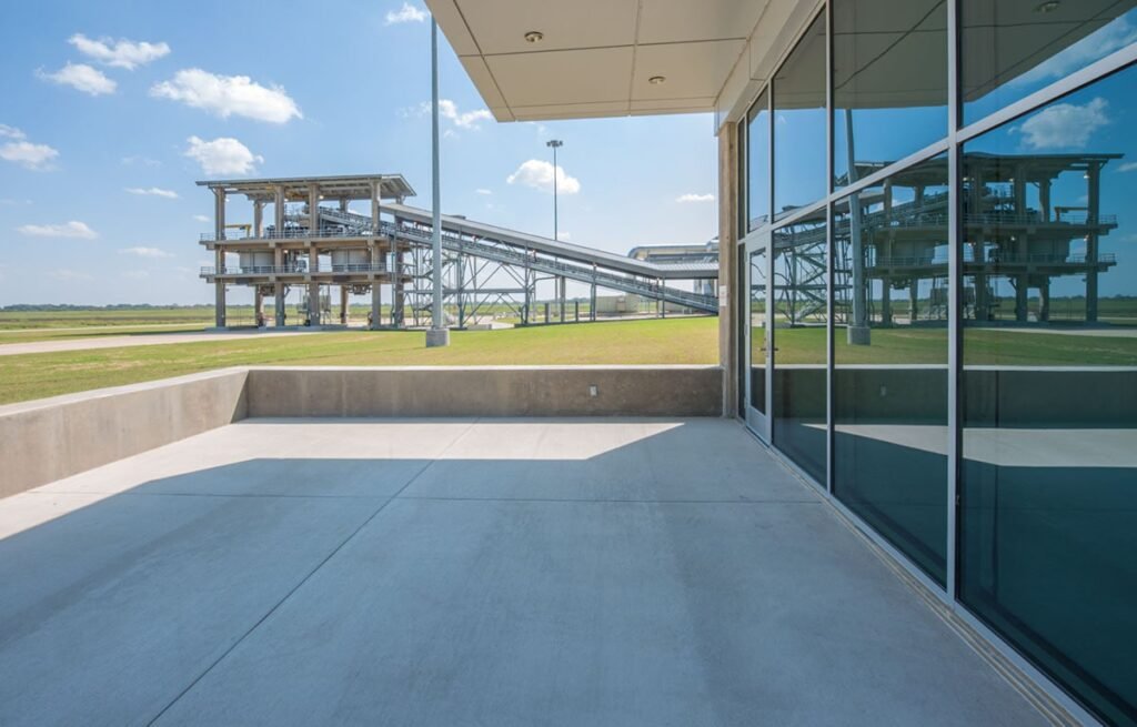 A modern building with large reflective glass windows gleams on a sunny day at the Dallas Treatment Plant. A concrete patio sits in the foreground, while metal industrial structures rise against a backdrop of clear blue sky and fluffy clouds.