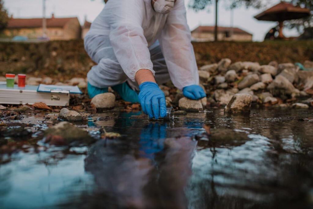 A person in protective clothing and gloves collects water in a vial from a rocky riverbed, with a test kit nearby, conducting an in-depth look into potential health risks. Trees and buildings are visible in the background.